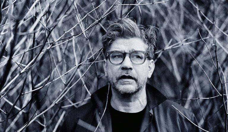 Zwart-witportret van een man met bril en baard, staand tussen hoog gras buiten.