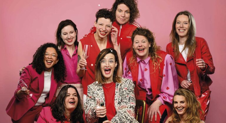 Een groep vrolijke mensen poseert tegen een roze achtergrond, gekleed in felgekleurde outfits.