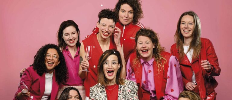 Een groep vrolijke mensen poseert tegen een roze achtergrond, gekleed in felgekleurde outfits.