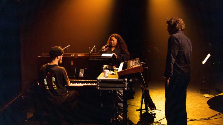 Drie muzikanten staan op een podium met een piano en synthesizer, onder een warme oranje verlichting.