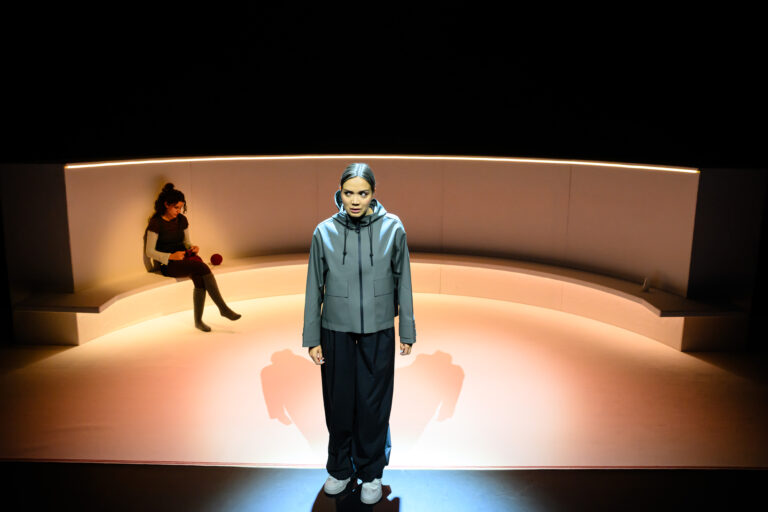 Een jonge vrouw staat in het midden van een podium, omgeven door zachte verlichting, terwijl een andere figuur op de achtergrond zit.