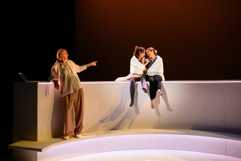 Een toneelscène met drie acteurs; twee zitten op een witte rand en fluisteren naar elkaar, terwijl een derde acteur achter hen staat en iets aanwijst.