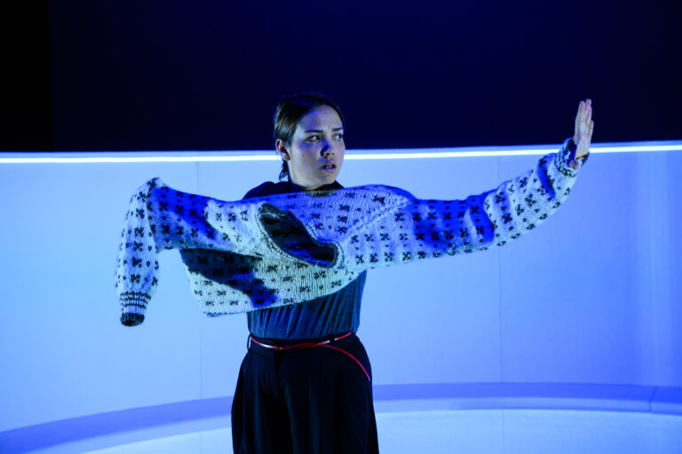 Een performer in een kleurrijke jurk met een opvallende schoudermantel, staande in een theateromgeving met blauw licht.