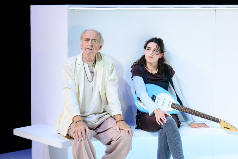 Een oudere man en een jonge vrouw zitten naast elkaar op een witte bank met een gitaren in de hand van de vrouw.