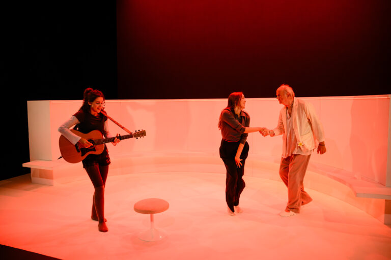 Twee acteurs in een theater omgeving met felrode verlichting, terwijl een derde acteur gitaar speelt.