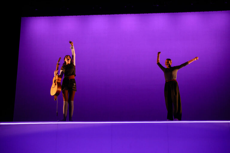 Twee performers op een podium met paarse achtergrond, één met een gitaar en de ander met uitgestrekte armen.