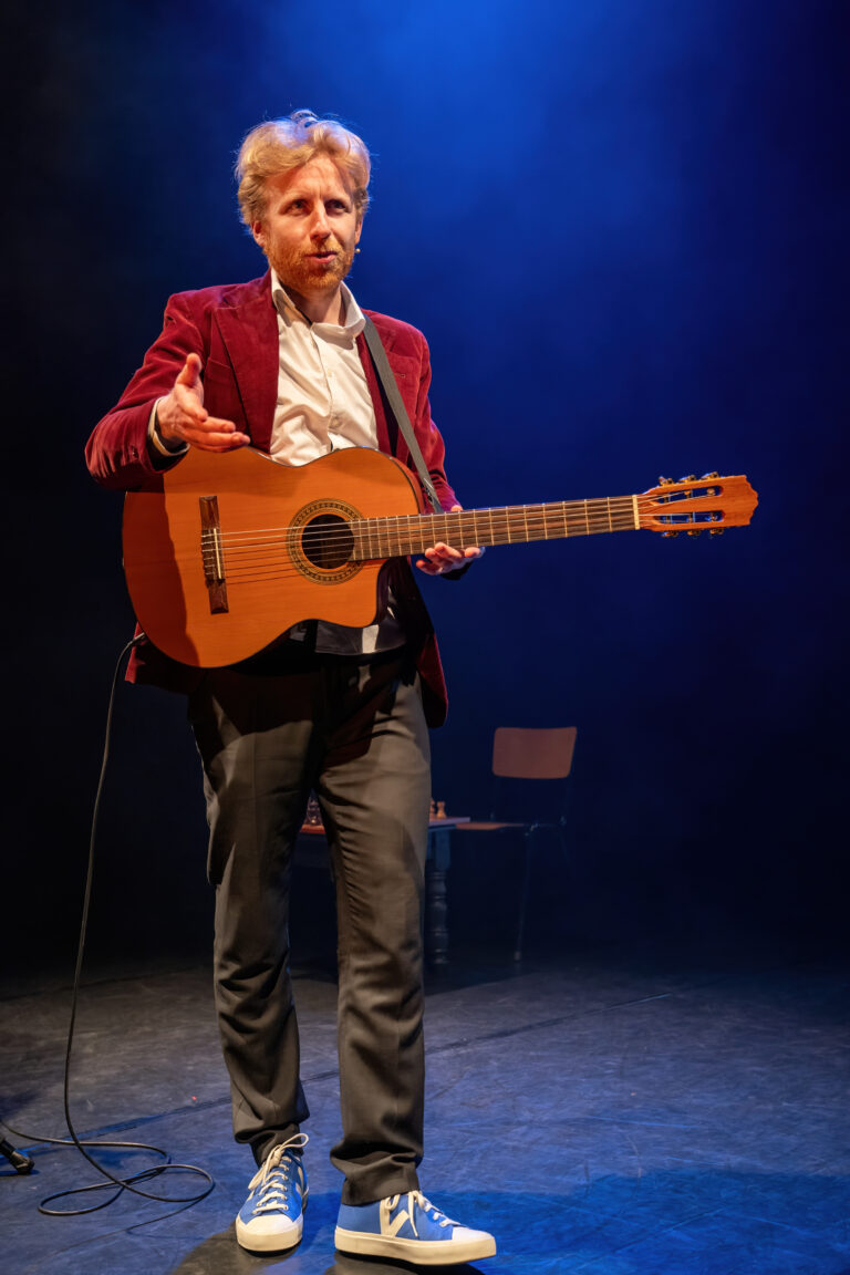 Een man in een rood jasje en blauwe sneakers staat op een podium met een gitaar, terwijl hij naar het publiek wijst.
