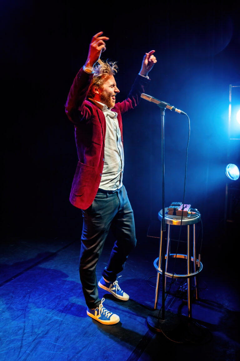 Een performer met een rood jasje staat op een podium en maakt gebaren terwijl hij een microfoon vasthoudt, omringd door blauwe verlichting.