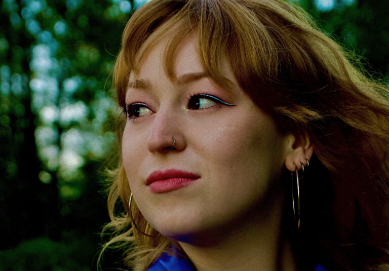 Close-up van een vrouw met lichtbruin haar en grote hoepeloorbellen die naar links kijkt tegen een groene buitenachtergrond.