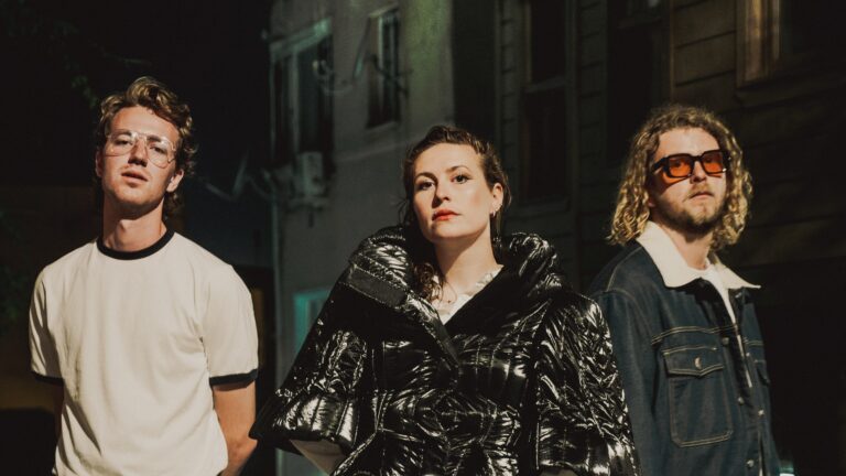 Een groep van drie mensen poseert buiten, gekleed in moderne, opvallende mode, met een stadsachtergrond in het donker.