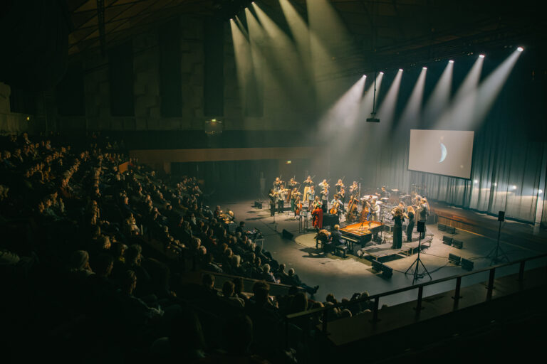 Een drukbezocht concert met een band op het podium, omringd door een enthousiaste menigte in een grote zaal.