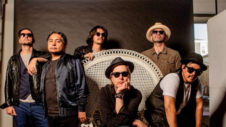 Chuck Prophet zit op een grote witte stoel met ronde hoge leuning omringt door zijn 5 bandleden.