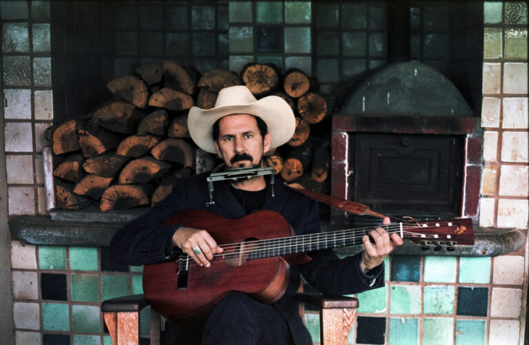 Een man in een cowboyhoed zit met een gitaar en een harmonica, met een achtergrond van gestapelde houtblokken.