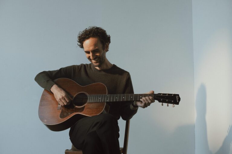 Een man speelt gitaar terwijl hij glimlacht, zittend op een stoel met een lichte achtergrond.