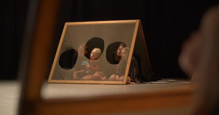 Drie kinderen zitten achter een houten scherm met doorzichtige gaten, op een podium.