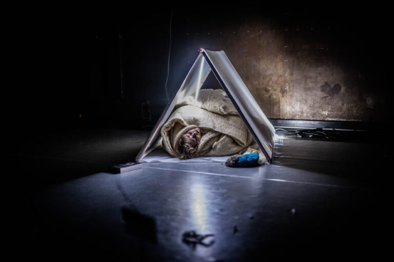 Een persoon ligt gewikkeld in dekens onder een omgekeerd boek als een tent in een donkere ruimte.