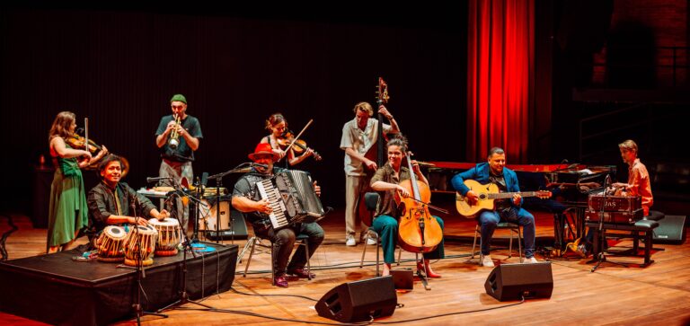 Een groep muzikanten speelt samen op een podium met verschillende instrumenten, waaronder een accordeon, cello en gitaar.