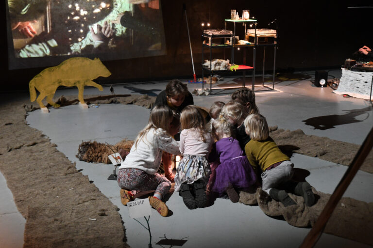 Een groep kinderen en een volwassene zit samen op de vloer van een creatieve ruimte, bezig met een activiteit rond een kunstinstallatie.