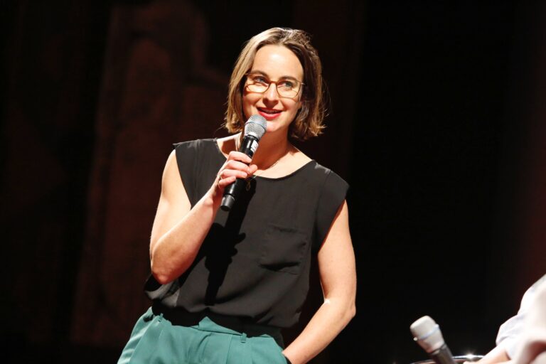 Een persoon met een microfoon staat op het podium en spreekt tijdens een evenement.