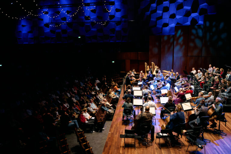Een orkest speelt voor een publiek in een sfeervolle concertzaal met blauwe verlichting.