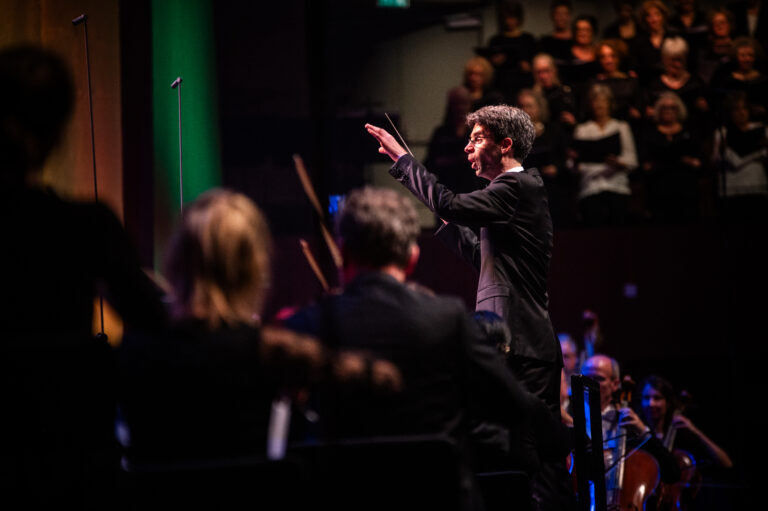 Een dirigent staat op het podium en dirigeert een orkest, terwijl het publiek in de achtergrond te zien is.