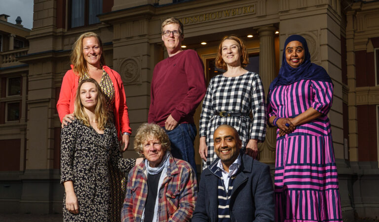 Een groep van zeven mensen poseert buiten een gebouw, gekleed in diverse outfits.