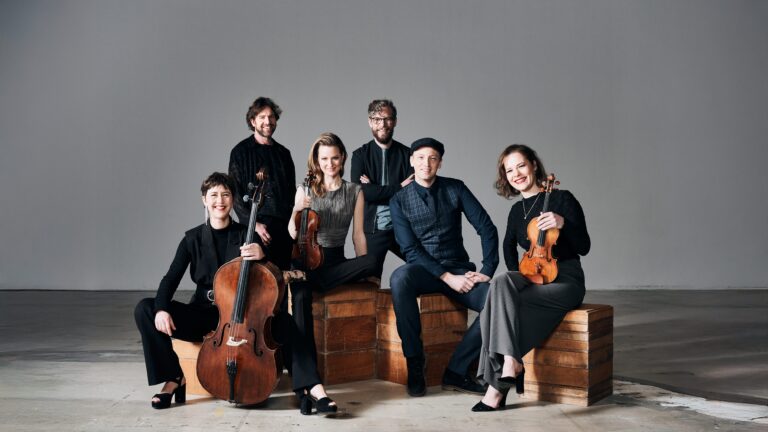 Zes muzikanten in zwarte kleding poseren samen op houten kratten in een moderne, minimalistische ruimte.