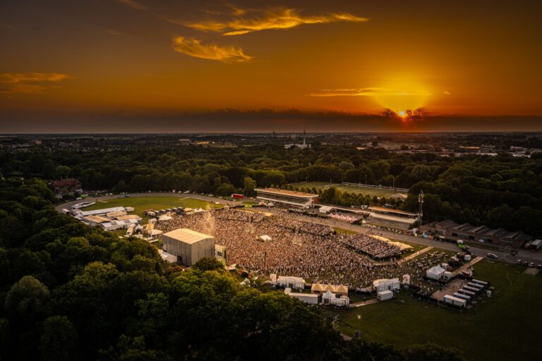 Een luchtfoto van een drukbezocht muziekfestival tijdens zonsondergang, met een groot aantal bezoekers en een prachtig uitzicht op de horizon.