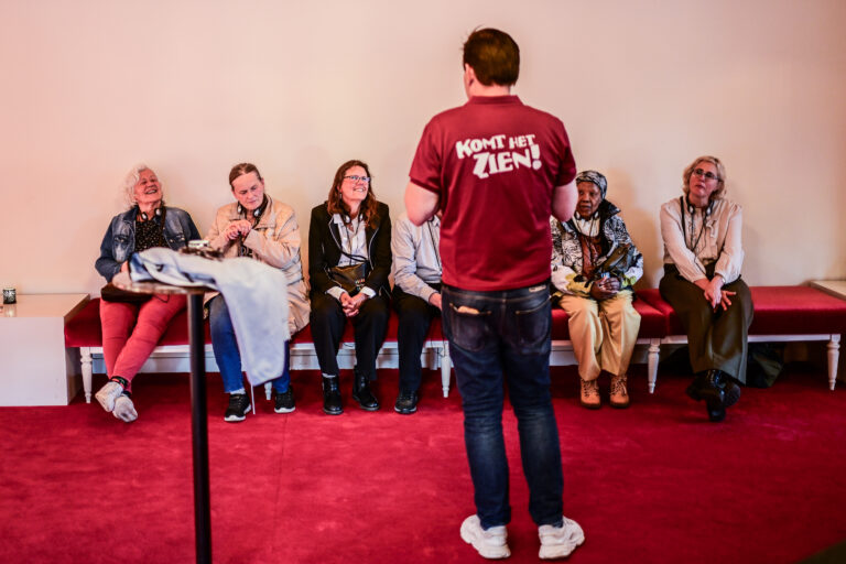 Een spreker staat met zijn rug naar het publiek in een zaal met een rode vloer, terwijl de aanwezigen aandachtig luisteren.