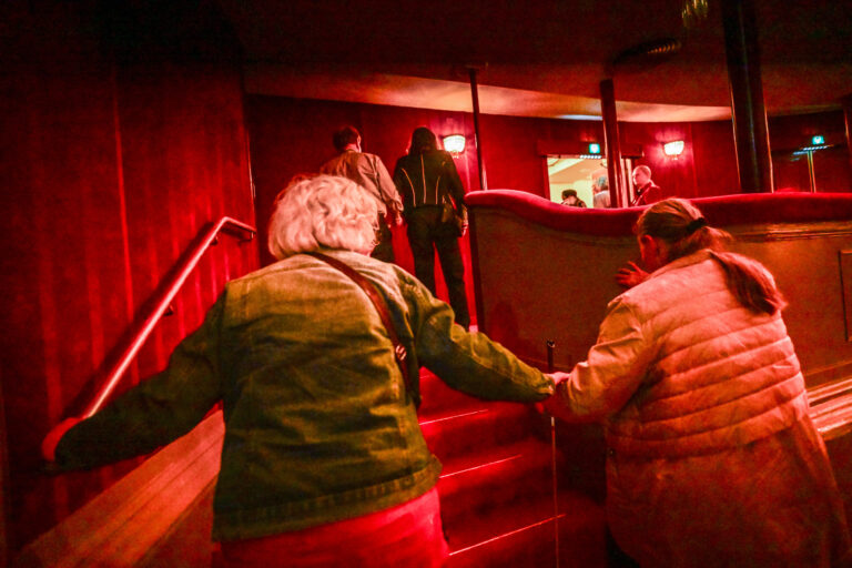 Oudere mensen lopen samen een trap op in een ruimte met een warme, rode verlichting.