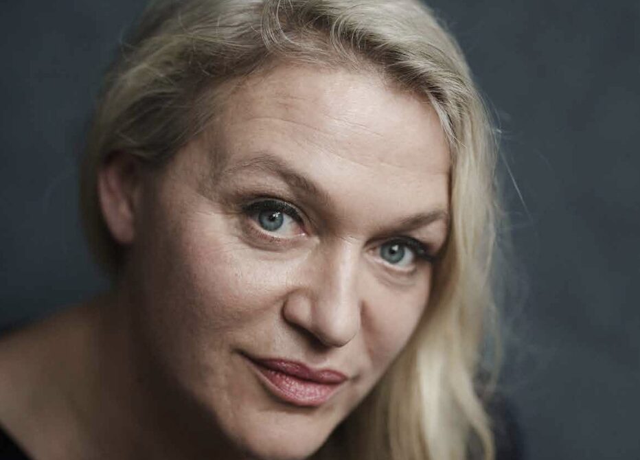 Een vrouw met lang, blond haar kijkt recht in de camera met een lichte glimlach.