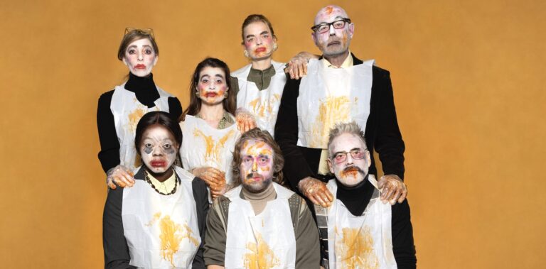Groep mensen in satirische kleding met opvallende gezichten en geschilderde make-up, poserend tegen een oranje achtergrond.