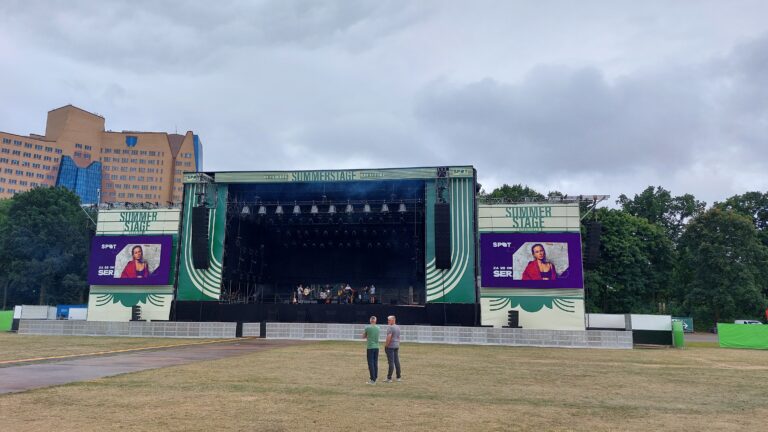 Een lege concertstage met een groot scherm, omringd door gras in een parkachtige omgeving.