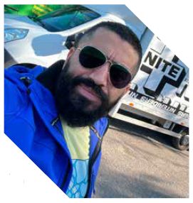 Een man met een baard en zonnebril poseert in een blauwe jas voor een witte vrachtwagen.