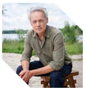 Een man in een groen overhemd zit op een houten kruk aan een waterkant, omringd door natuur.