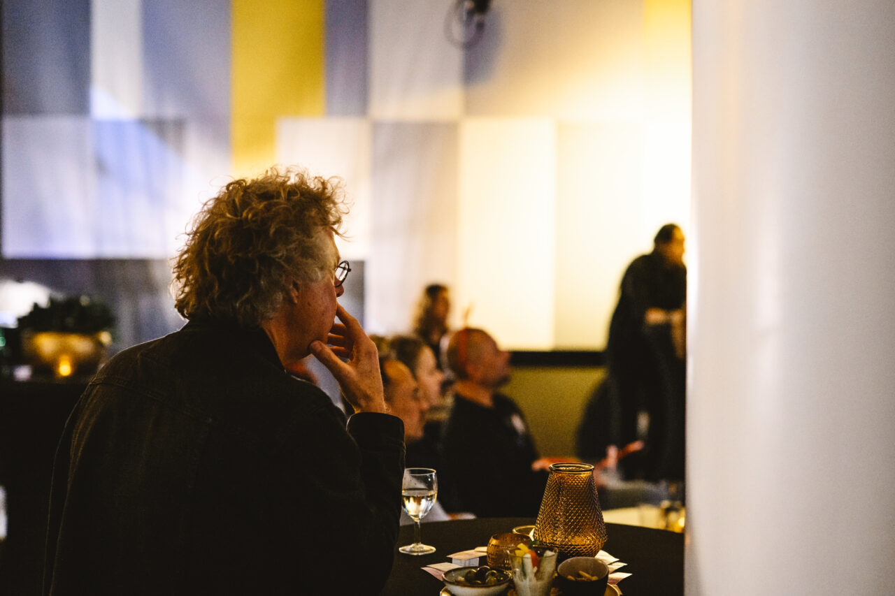 Man met krullend grijs haar kijkt geconcentreerd richting een podium of presentatie, zittend aan een tafel met een glas water en snacks.