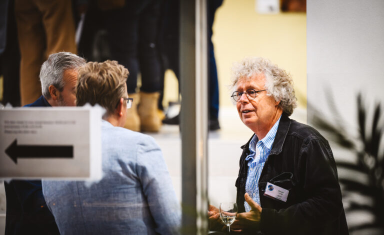 Man met krullend grijs haar en bril voert een gesprek met twee andere mensen tijdens een netwerkborrel; hij houdt een glas vast.