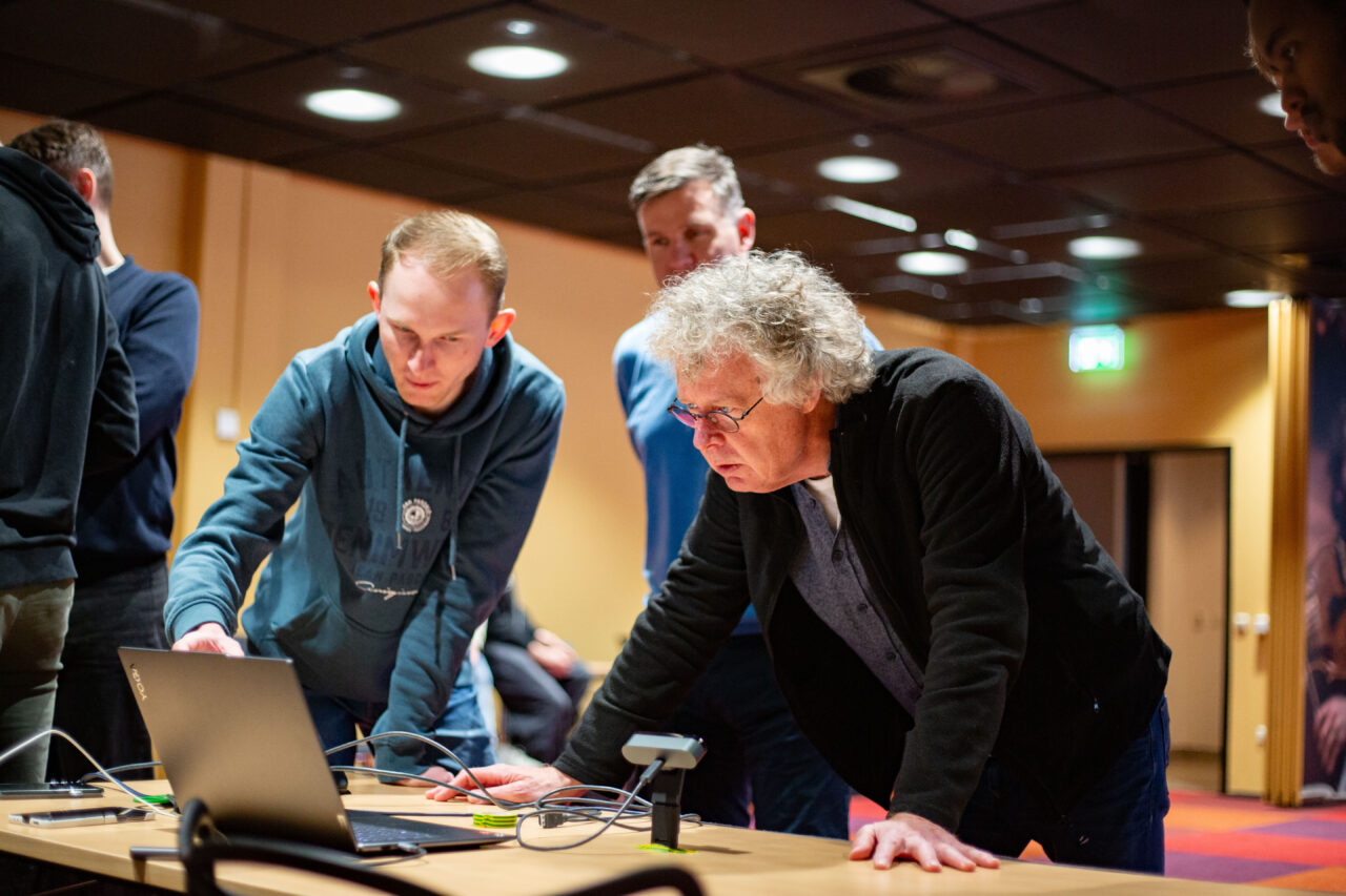 Man met krullend grijs haar buigt voorover naast een jonge man in hoodie, terwijl ze samen naar een laptop kijken tijdens een workshop of technische sessie.