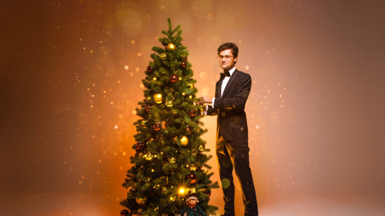 Een man in een tuxedo staat naast een prachtig versierde kerstboom met een geschenk.