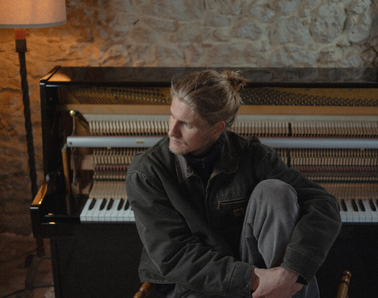 Man met een bruine jas en bruine broek staart voor zich uit en zit op een stoel voor de piano.