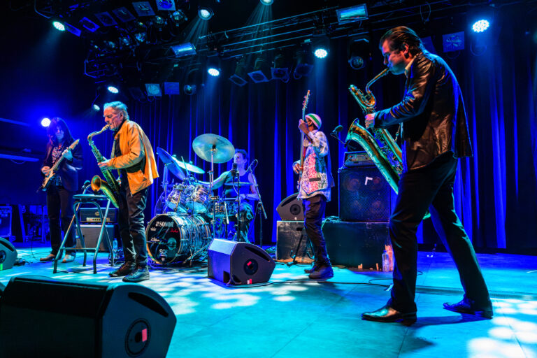 Vier mannen op een blauw belicht podium met saxofoon, drum en gitaar.