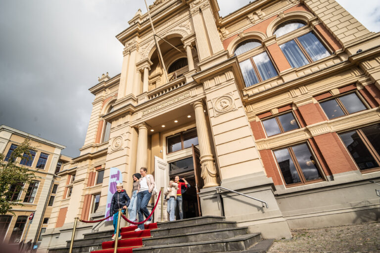 Een groep mensen staat bij de ingang van een elegant gebouw met grote ramen en een trap, afgewerkt met een rode loper.