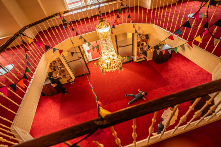 Een bovenaanzicht van een ruime ingang met een rode vloerbedekking, een grote kroonluchter in het midden en feestelijke vlaggetjes aan de leuning.