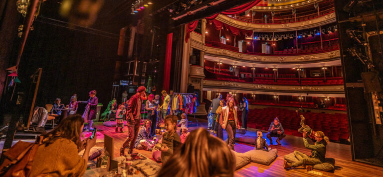 Een levendig theater met een variété aan kunstenaars die zich voorbereiden op een optreden, terwijl het publiek in de achtergrond zit.