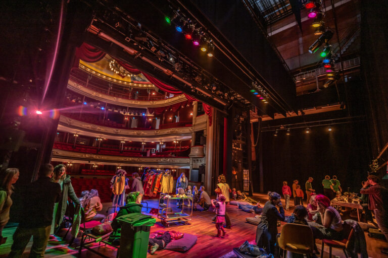 Een druk theater achter het podium met acteurs en crewleden die zich voorbereiden, omringd door kleurrijke lichten.