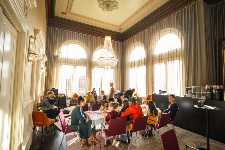 Een gezellige ruimte met grote ramen en een kroonluchter, waar een groep mensen aan tafels zit.