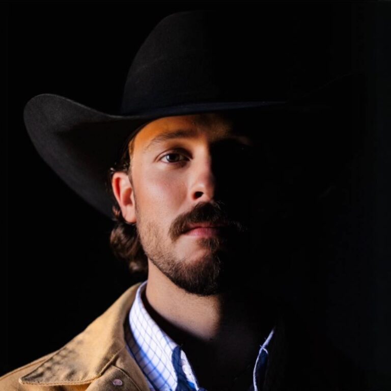 Een man met een hoed kijkt serieus naar de camera, terwijl zijn gezicht voor een deel in de schaduw valt.