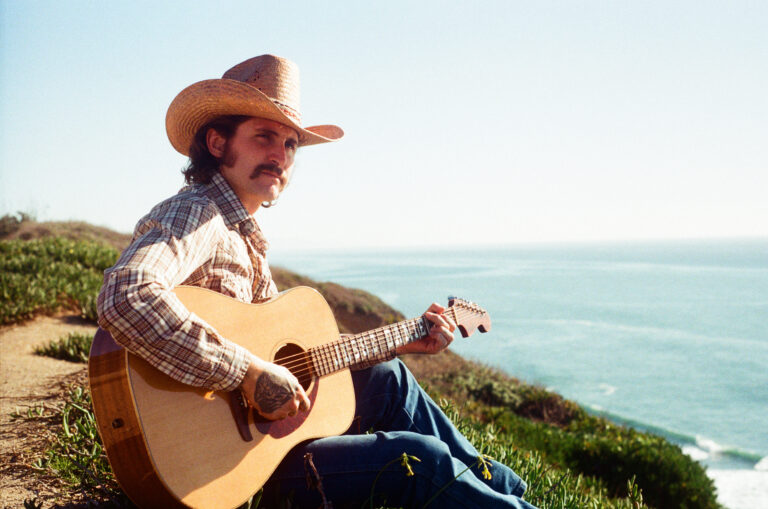 Een man in een cowboyhoed speelt gitaar aan de rand van een kustlijn met de zee op de achtergrond.