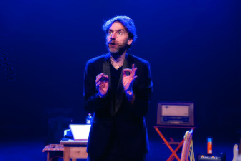 Een man staat op het podium met een expressieve uitstraling, gefocust op zijn handen terwijl hij interactie heeft met het publiek.