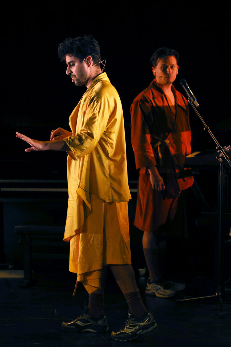 Acteur in geel gewaad kijkt naar zijn hand, met acteur in bruin gewaad die naar hem kijkt